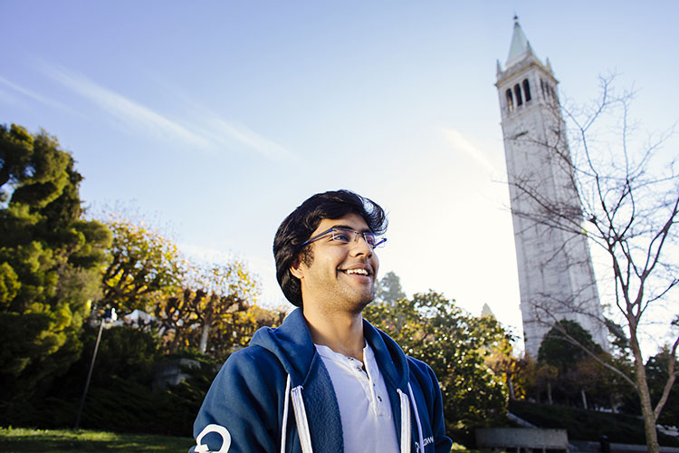 A young man walks across campus on a sunny day with clear blue skies while the Campanile towers high in the background. 