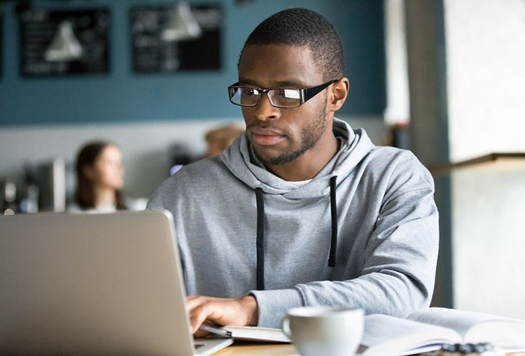 A young, black male wearing eye glasses and a gray hoodie studies at a cafe using his laptop, books, and notebooks. 