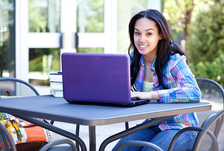  A student siding outside at a table smiling typing on a laptop.