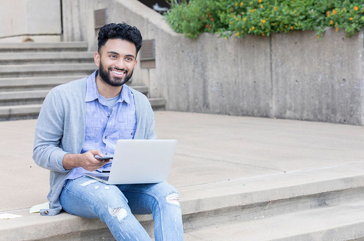  A student sitting on the steps of a building holding their laptop and phone.