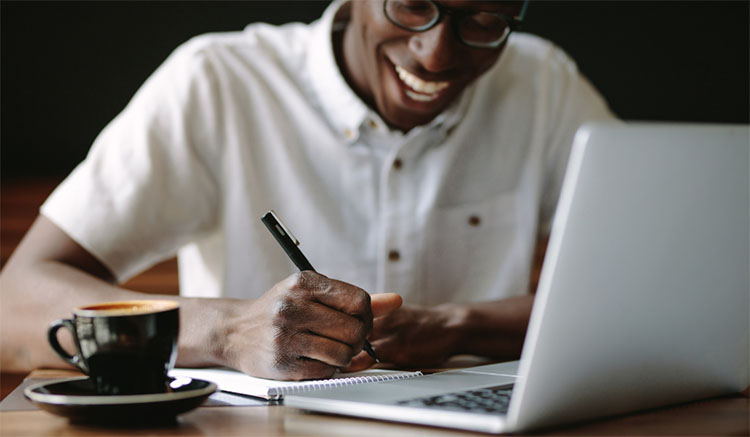  A student smiling while taking notes in front of a laptop and a cup of coffee.