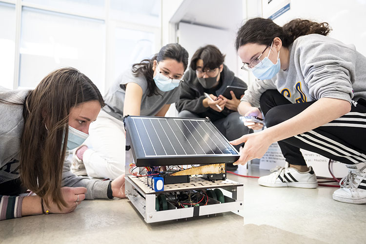 Three female students and one male student, all dressed in casual clothing, sneakers, and Cal hoodies, kneel down to work on a technical device that is sitting on the floor in front of them. 