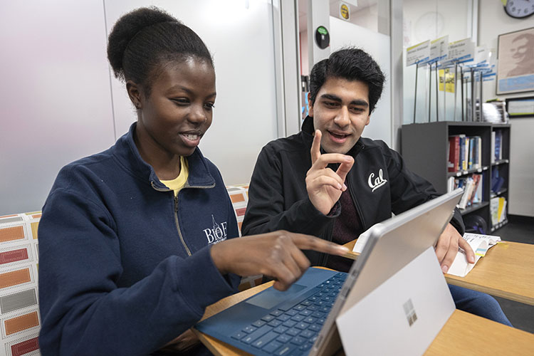 A young, black, female student sits next to a male student with dark hair. Each are wearing Cal gear. They are sitting a study space on campus with a bookshelf behind them, and a book and a laptop in front of them. 