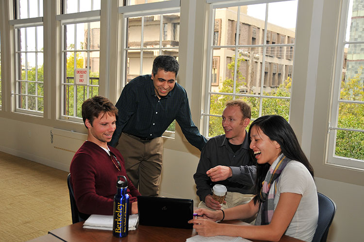 A male professional is standing and leaning into a conversation between three young adults who are sitting at a table in an office. They are discussing something that they see on the screen of a laptop. One of the men is pointing at the screen. They are all smiling and laughing. There is a row of tall windows behind them with a view of bushes and another large building in the background. 