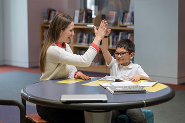 A teacher and a young student sitting at a round table giving each other a high-five.